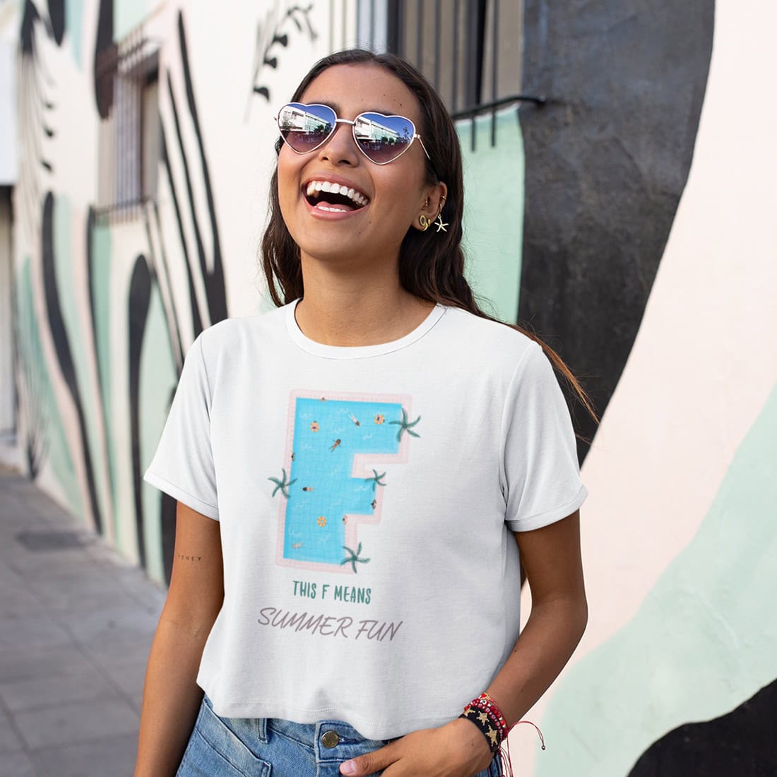 Junge Frau mit dunklem Haar und Herzsonnenbrille steht lachend vor Graffitti-Hauswand. Sie trägt weißes T-Shirt mit Print.