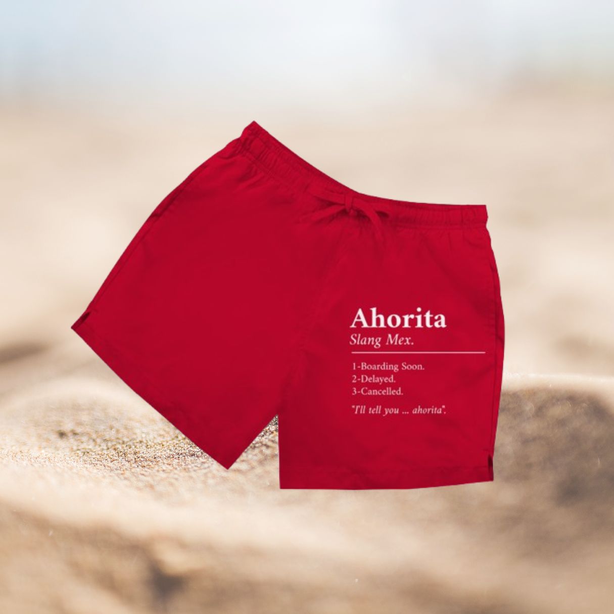 Rote Herren Badeshorts mit Ahorita (Slang Mex) Aufdruck vor Sandhintergrund.