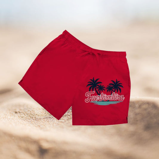 Rote Herren Badeshorts mit Palmenmotiv und Schriftzug Fuerteventura vor Sandhintergrund.