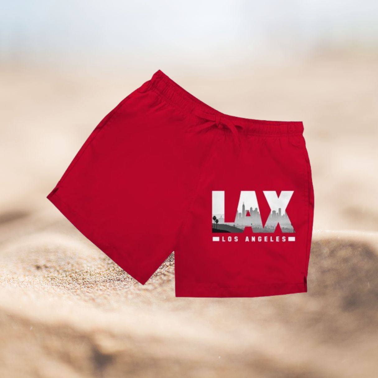 Rote Herren Badeshorts mit Aufdruck "LAX" (Airport Code) vor Sandhintergrund.