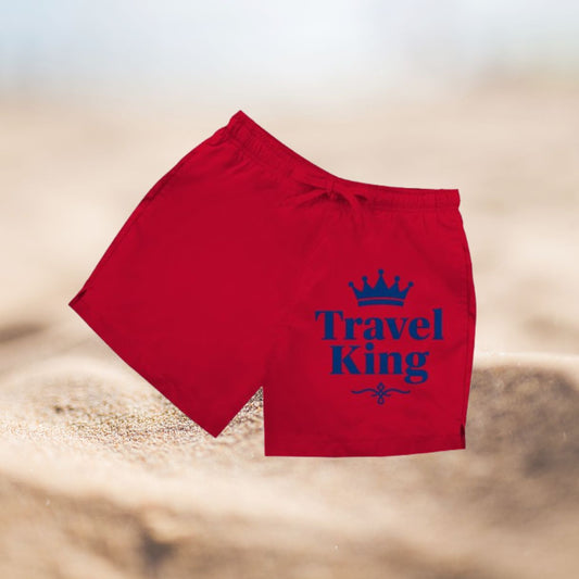 Rote Herren Badeshorts mit blauem Travel King Schriftzug vor Sandhintergrund.