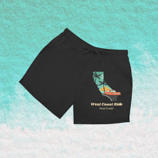 Schwarze Herren Badeshorts mit Schriftzug „West Coast Ride“ vor türkisfarbenem Wasserhintergrund.