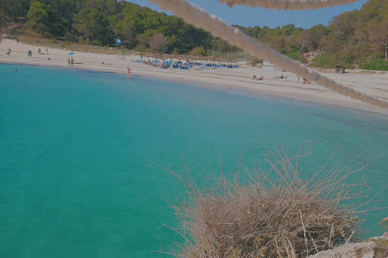 Türkisfarbenes Wasser mit einem breiten Sandstrand mit einigen Leuten am und im Wasser (Playa de S'Amarador)