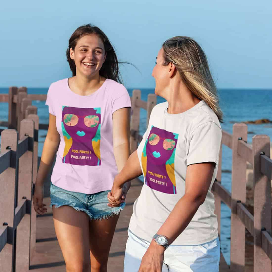 Zwei Mädchen Hand in Hand auf einer Holzbrücke am Meer. Sie tragen T-Shirts mit POOL PARTY Design in White und Orchid Pink.