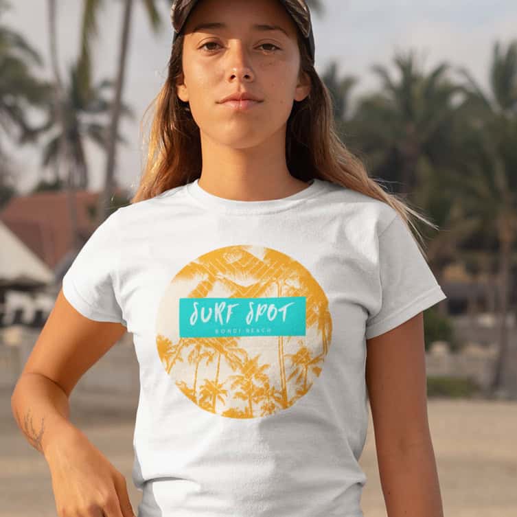 Junge Frau am Palmenstrand blickt in die Kamera, trägt weißes TShirt mit sandfarbenem Palmendesign und grünen Akzenten.