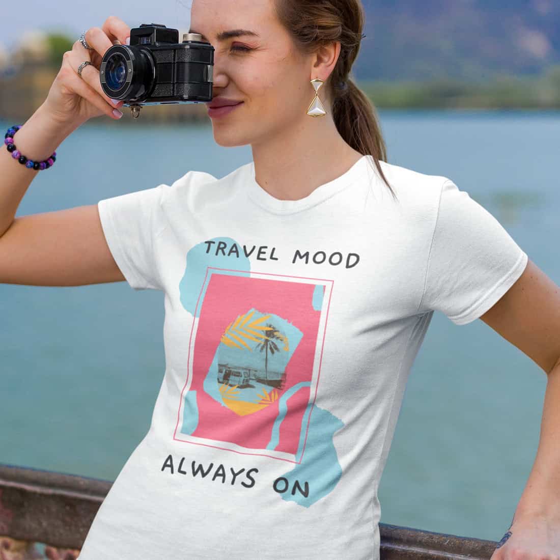 Junge Frau vor Berg und See-Kulisse mit Fotoapparat vorm Gesicht. Sie lehnt an Brüstung und trägt weißes T-Shirt mit Design.