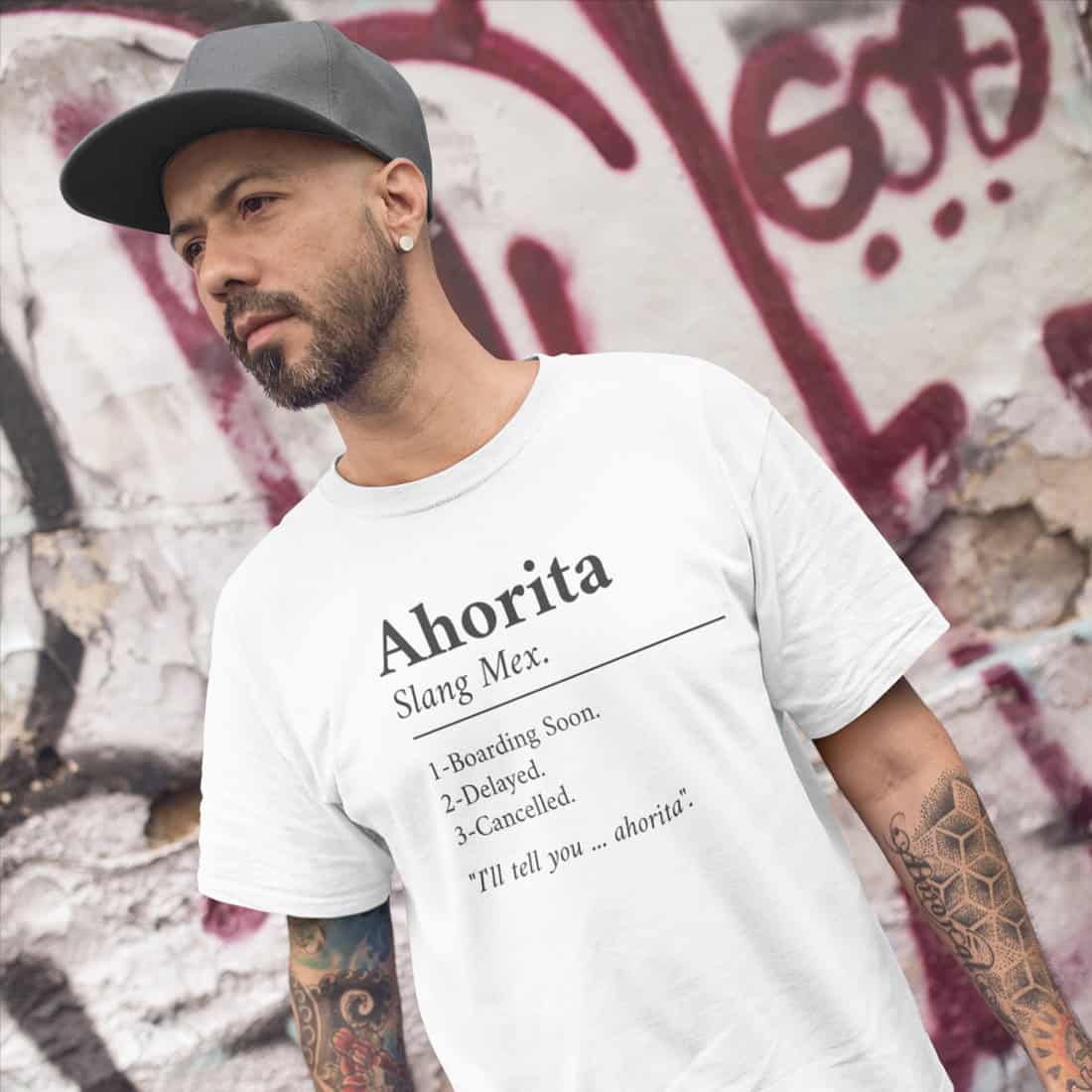 Junger tätowierter Mann mit Vollbart steht vor einer weißen Wand mit rotem Graffitti. Er trägt ein weißes T-Shirt mit Print.