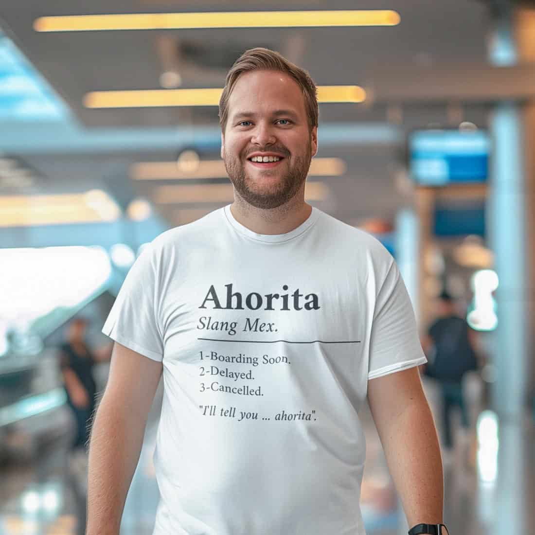 Kräftiger junger Mann mit Vollbart. Hintergrund unscharf, evtl. Flughafen. Er trägt ein weißes T-Shirt mit dem Design.