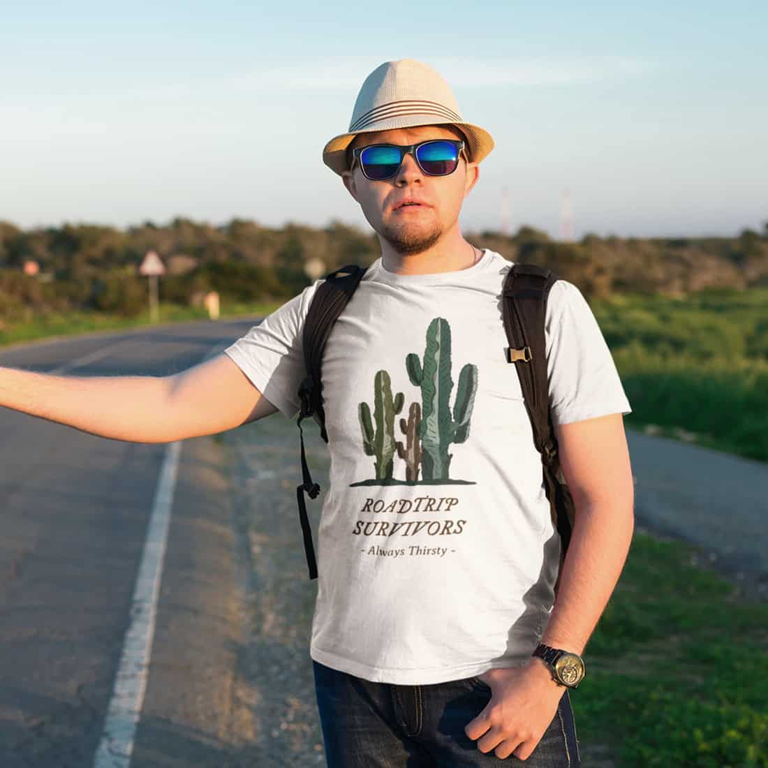 An einer Straße steht ein junger Mann mit Sonnenhut, Sonnenbrille und Rucksack. Daumen ausgestreckt. T-Shirt mit Design.