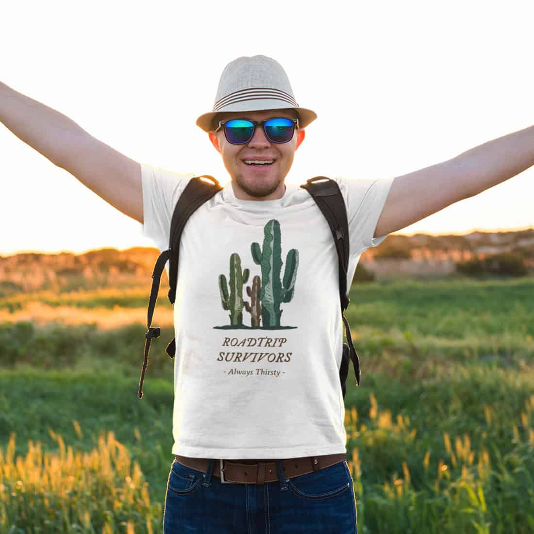 Mann mit Sonnenhut und Sonnenbrille steht mit ausgebreiteten Armen auf einer Wiese. Er trägt ein helles T-Shirt mit Print.