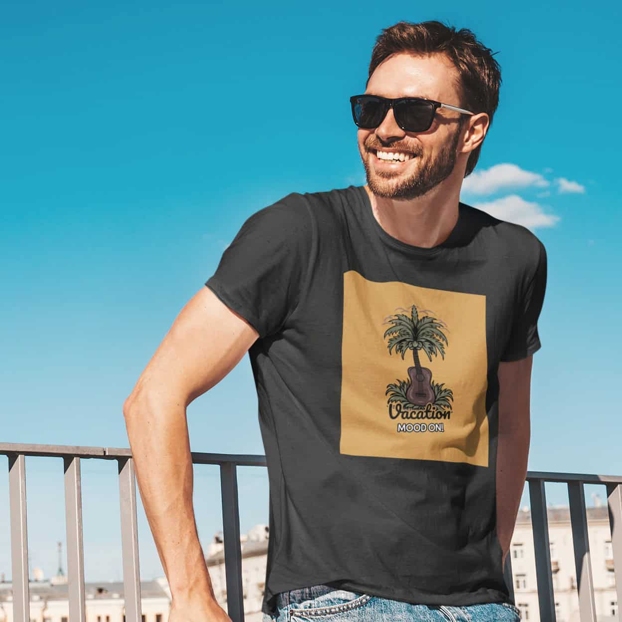 Blauer Himmel, im Hintergrund eine Stadt. Junger Mann lehnt an Geländer. Sonnenbrille, Lächeln.Schwarzes Tee mit gelbem Print