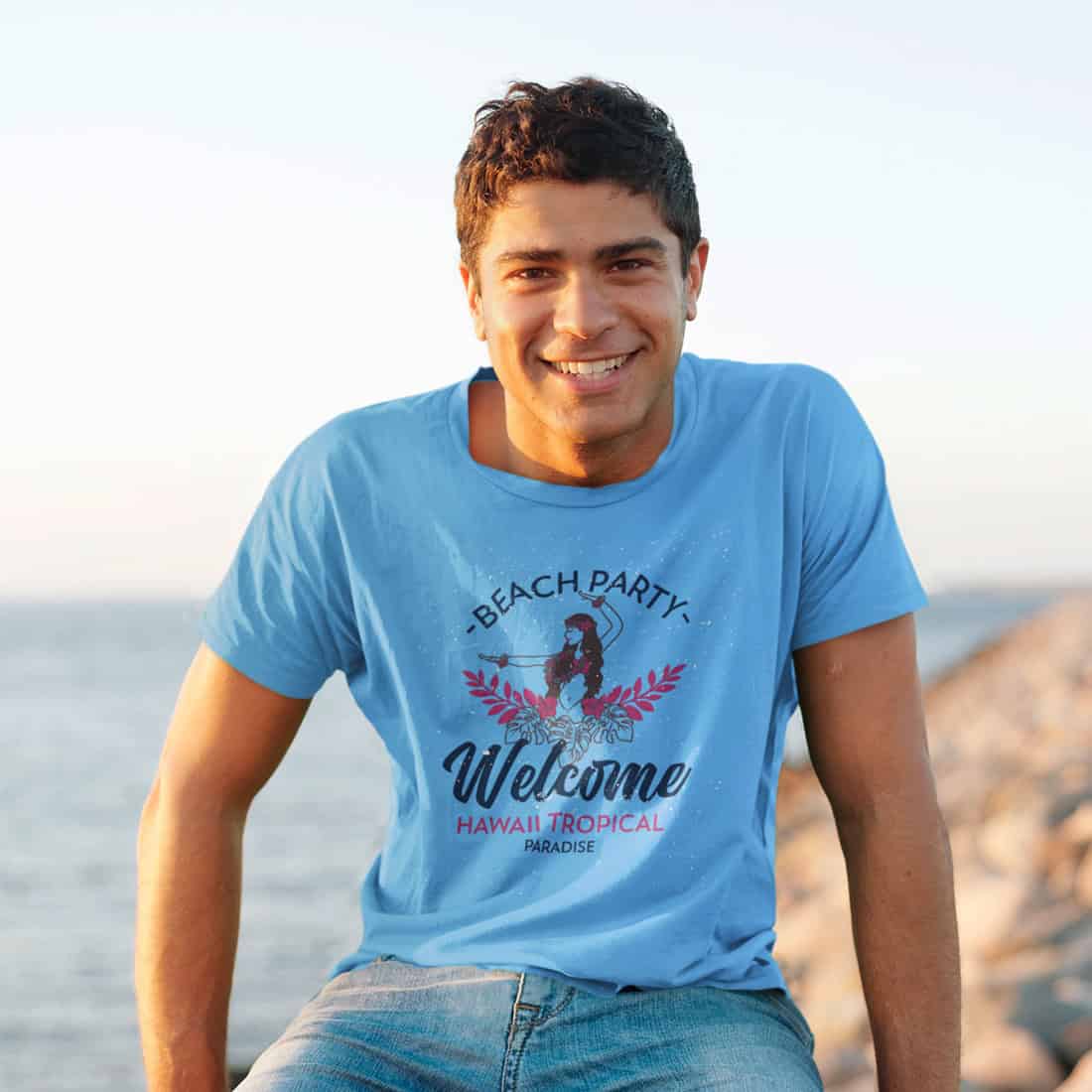 Lächelnder junger Mann auf einer Barke sitzend vor Meer und Steinküste. Er trägt hellblaues TShirt mit Design.