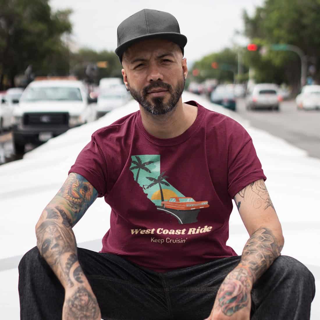 Lässiger Typ mit Basecap sitzt auf einem Dach. Er trägt schwarze Jeans und ein dunkelrots Shirt mit West Coast Ride Design.