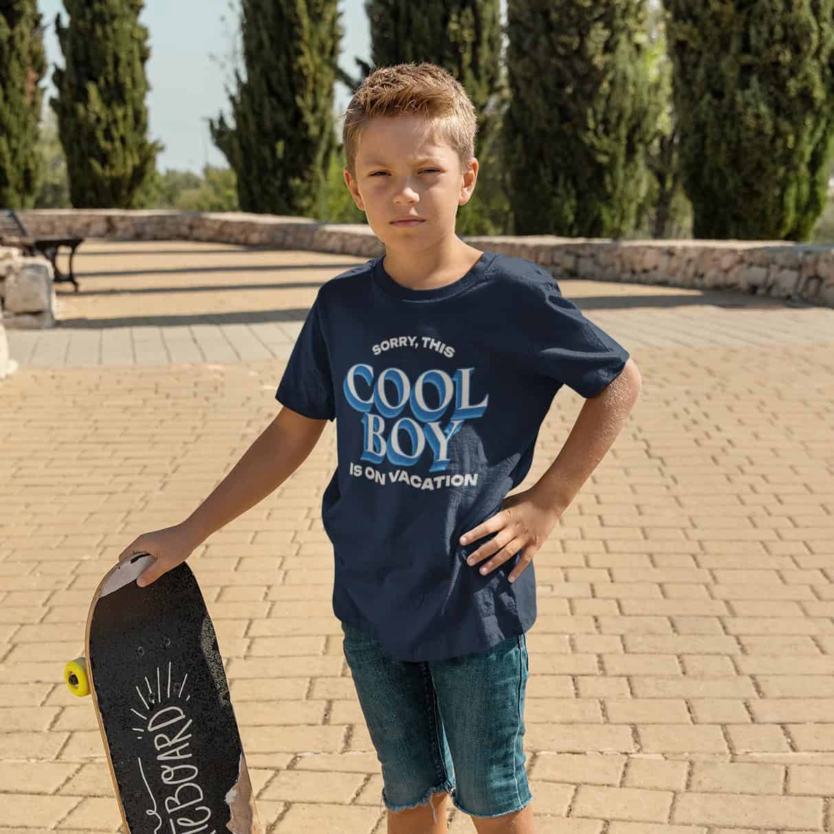 Junge mit Skateboard, kurzen Hosen und ernstem Blick trägt ein dunkelblaues T-Shirt dem Design-Namen.