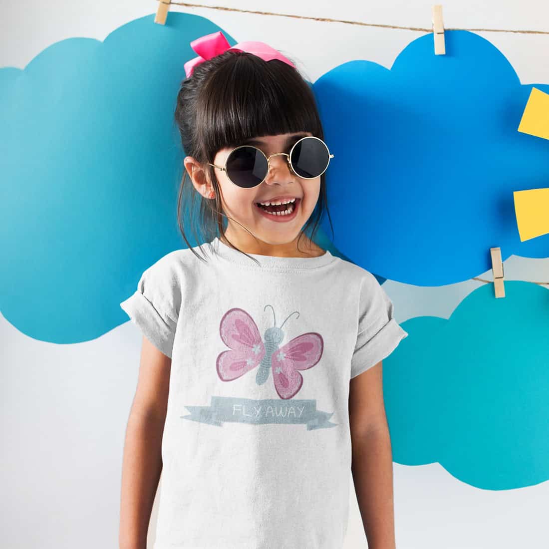 Kleines Mädchen mit dunklem Haar, Pony und pinkfarbener Schleife lacht in die Kamera. Sie trägt ein weißes T-Shirt mit Print.