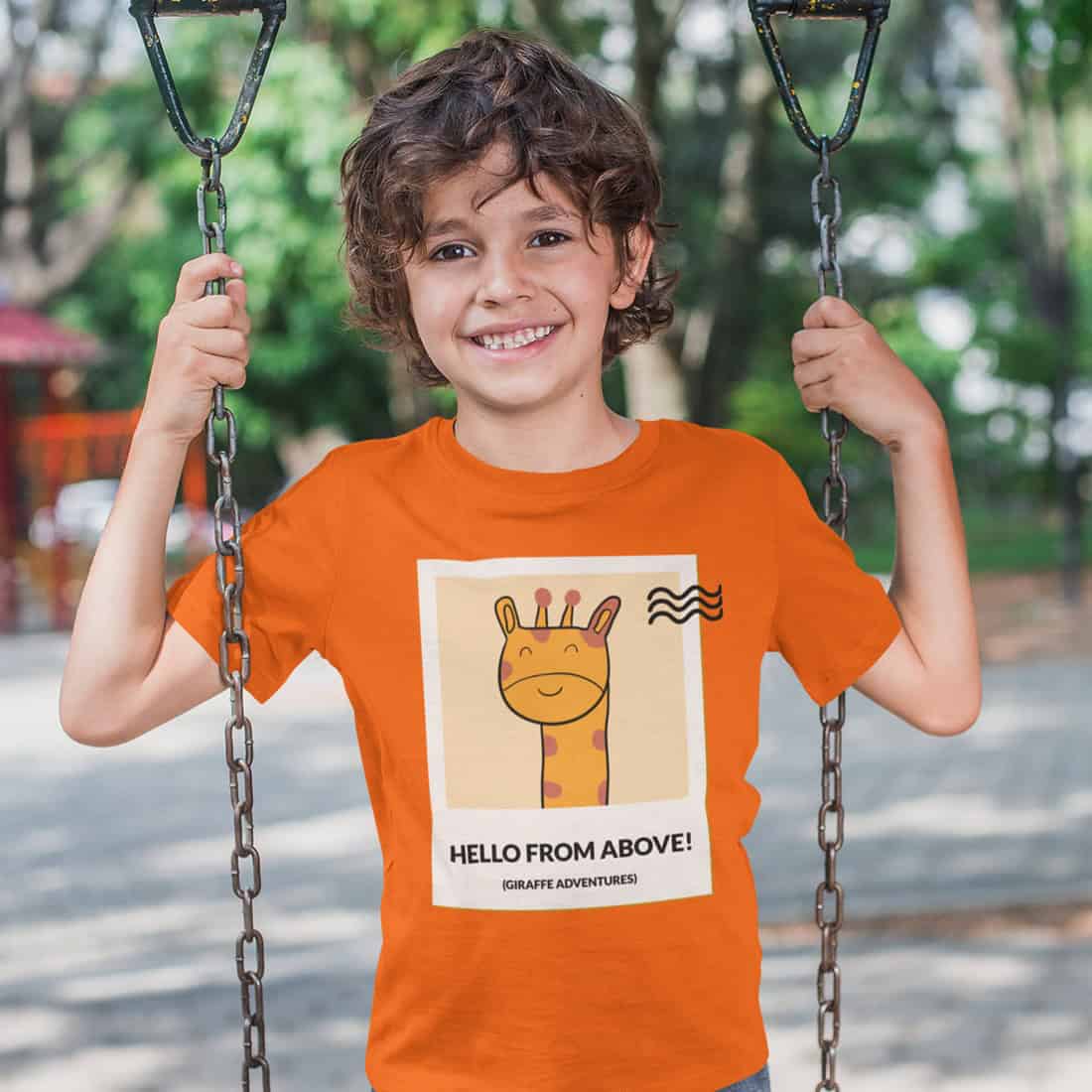 Im Stehen Schaukelnder Junge trägt ein orangefarbenes T-Shirt mit dem Design als Print. Er lächelt in die Kamera.