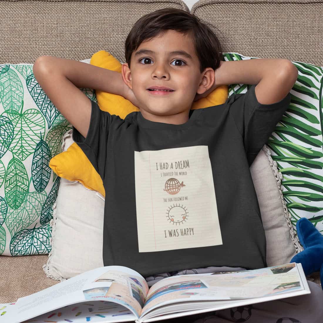 Junge lümmelt auf der Couch, ein Bilderbuch auf den Beinen. Er trägt ein schwarzes T-Shirt auf dem das Design gedruckt ist.