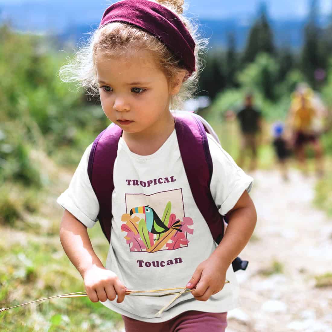 Kleines Mädchen mit lila Kopfband, Rucksack und Hose trägt ein weißes Shirt mit Toucan Print. Zwischen den Händen ein Zweig.
